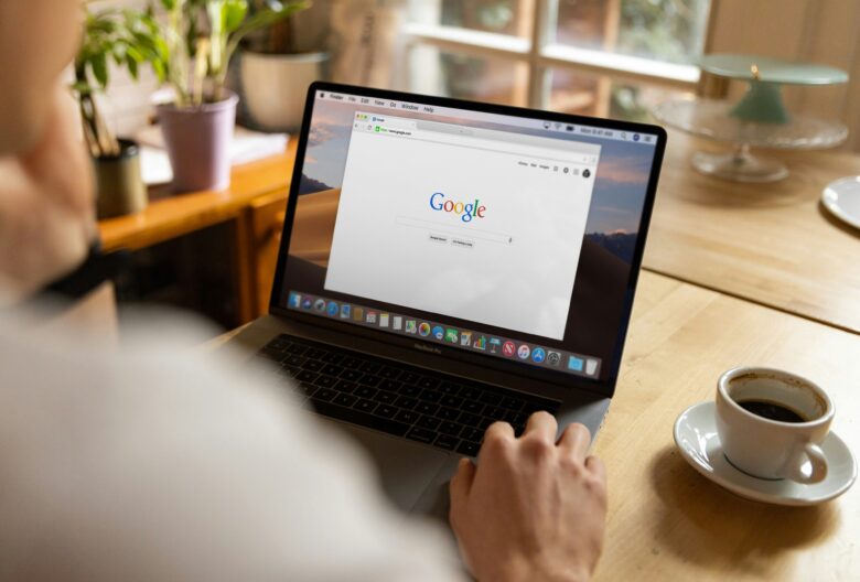 A person sitting at a wooden table using a MacBook Pro laptop with the Google homepage open on the screen. A cup of coffee sits beside the laptop, and potted plants and a window are visible in the background.