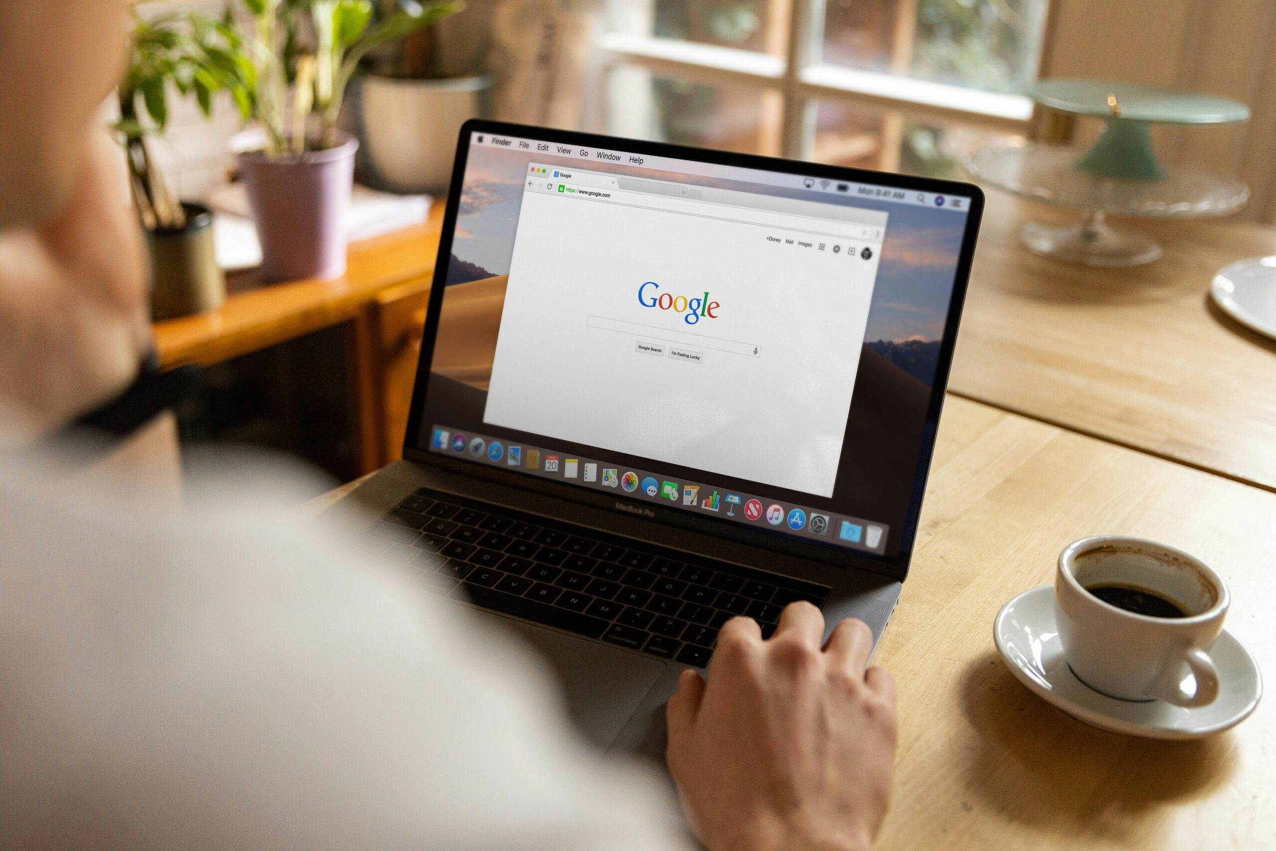 A person sitting at a wooden table using a MacBook Pro laptop with the Google homepage open on the screen. A cup of coffee sits beside the laptop, and potted plants and a window are visible in the background.
