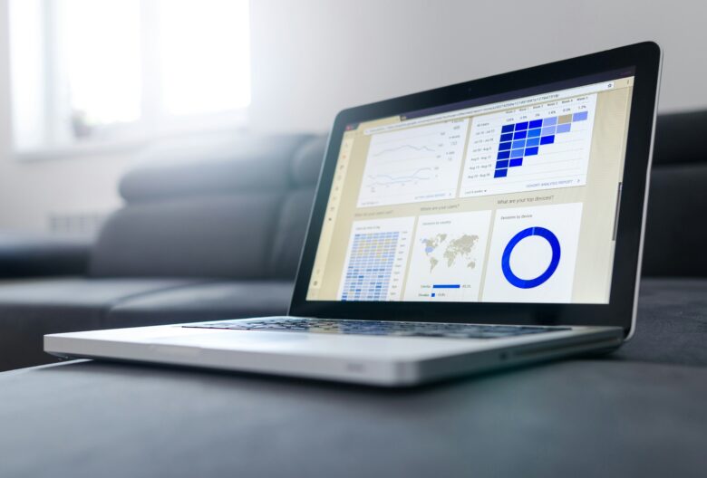A silver laptop on a dark grey sofa displaying a data analytics dashboard. The screen shows charts and graphs including a line graph, a heatmap, a world map highlighting countries, and a blue circular chart labeled “Sessions by device.” The room is bright with natural light coming through a window in the background.