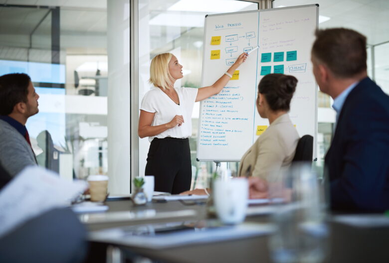 Marketing team gathered in a meeting as a presenter explains a web strategy process on a whiteboard during a collaborative planning session.